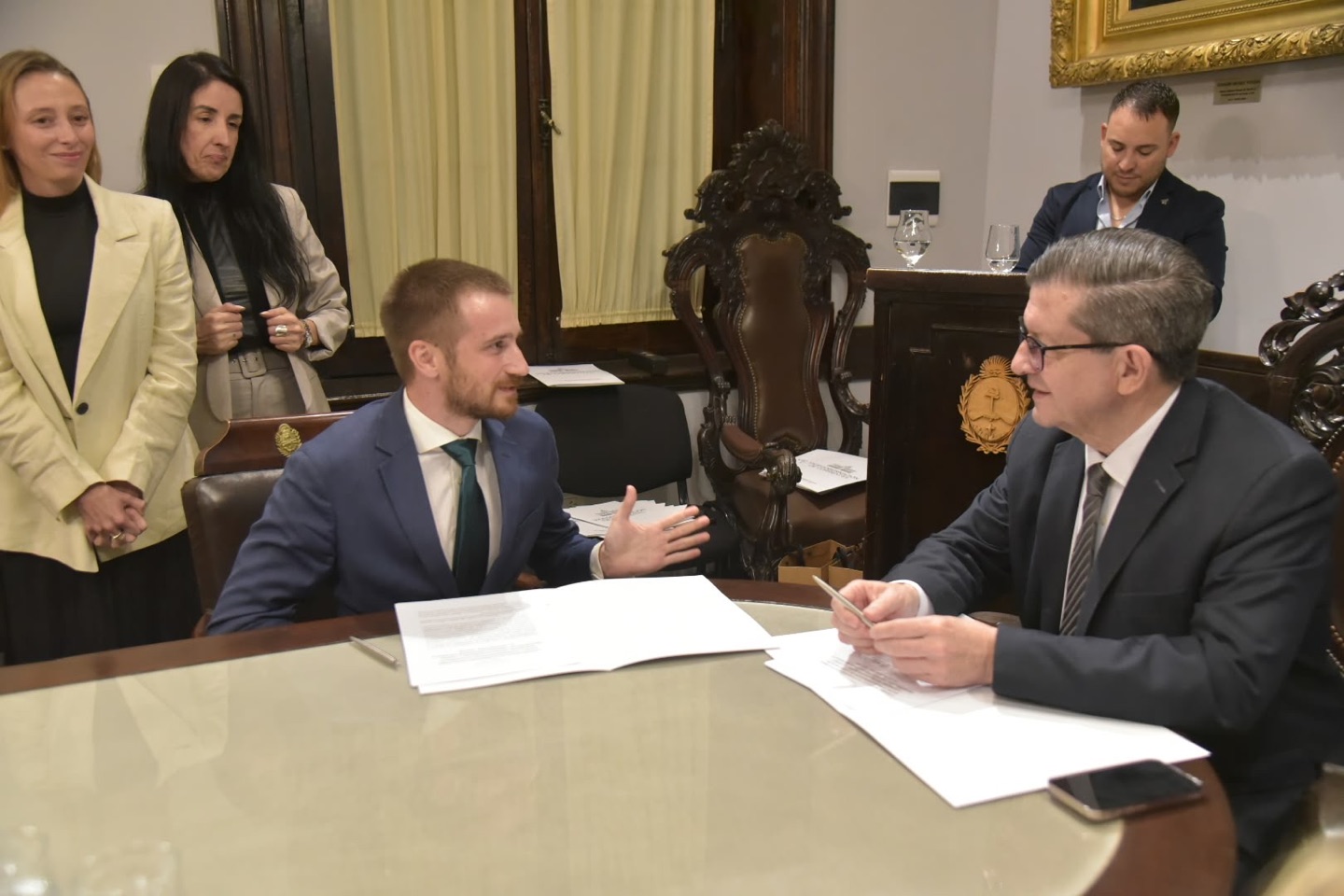 Jakob Saalfrank, seated at the signing table, explains the agreement to Vice-Governor Néstor Pedro Braillard Poccard, with the papers between them and others standing behind.